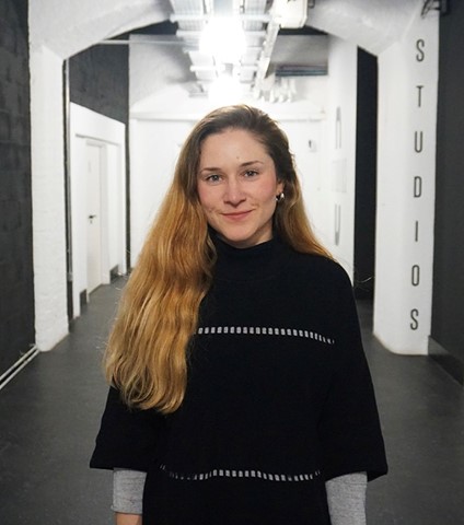 A young female adult with long, wavy blone/brown hair, wearing a black jumper, standing in a corridor with charcoal flooring, black walls, white columns and archs and door to the right and behind.
